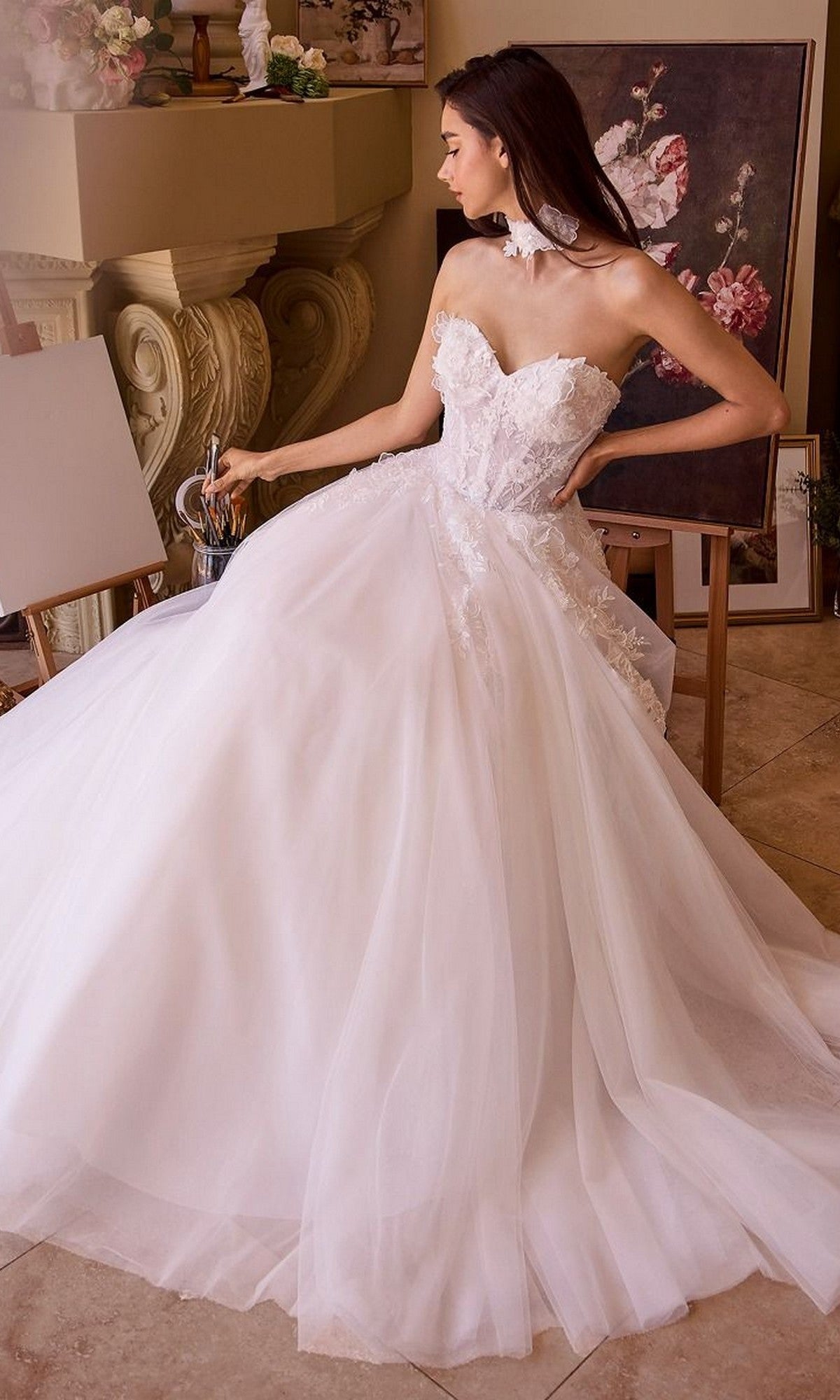 A woman wearing the Andrea and Leo WL041 long wedding dress with a strapless floral lace bodice sits indoors near artwork, holding a clear glass pitcher and looking to her left.